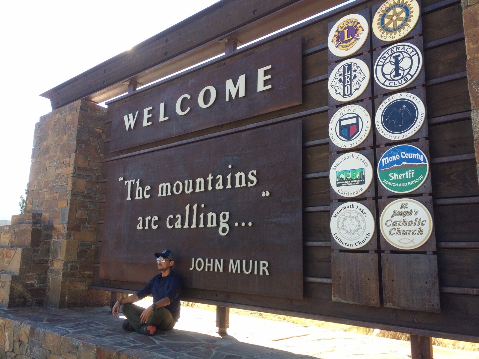 Welcome to Mammoth Hero image of Isaac Medrano sitting in front of the Welcome to Mammoth sign. "The mountain are calling..." John Muir quote.
